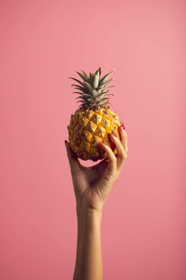 A hand with polished nails holds a ripe pineapple against a vivid pink backdrop, embodying tropical freshness.